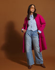 Woman wearing the orchid coat, light blue jeans, and sunglasses on a brown background.
