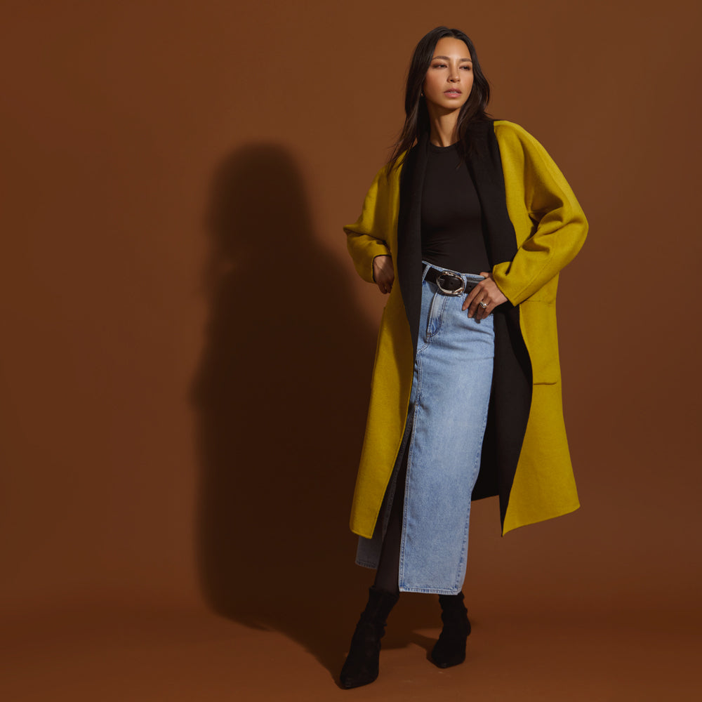 Woman wearing a moss and black coat with a black top, and blue jeans against a brown background.