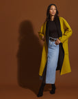 Woman wearing a moss and black coat with a black top, and blue jeans against a brown background.
