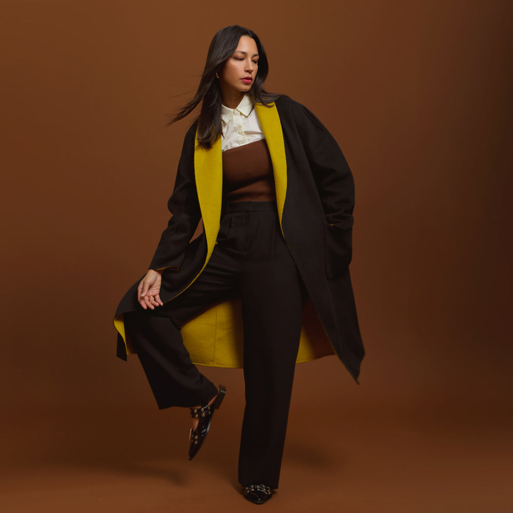 Woman in a stylish outfit with a moss and black coat against a brown background.