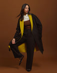 Woman in a stylish outfit with a moss and black coat against a brown background.