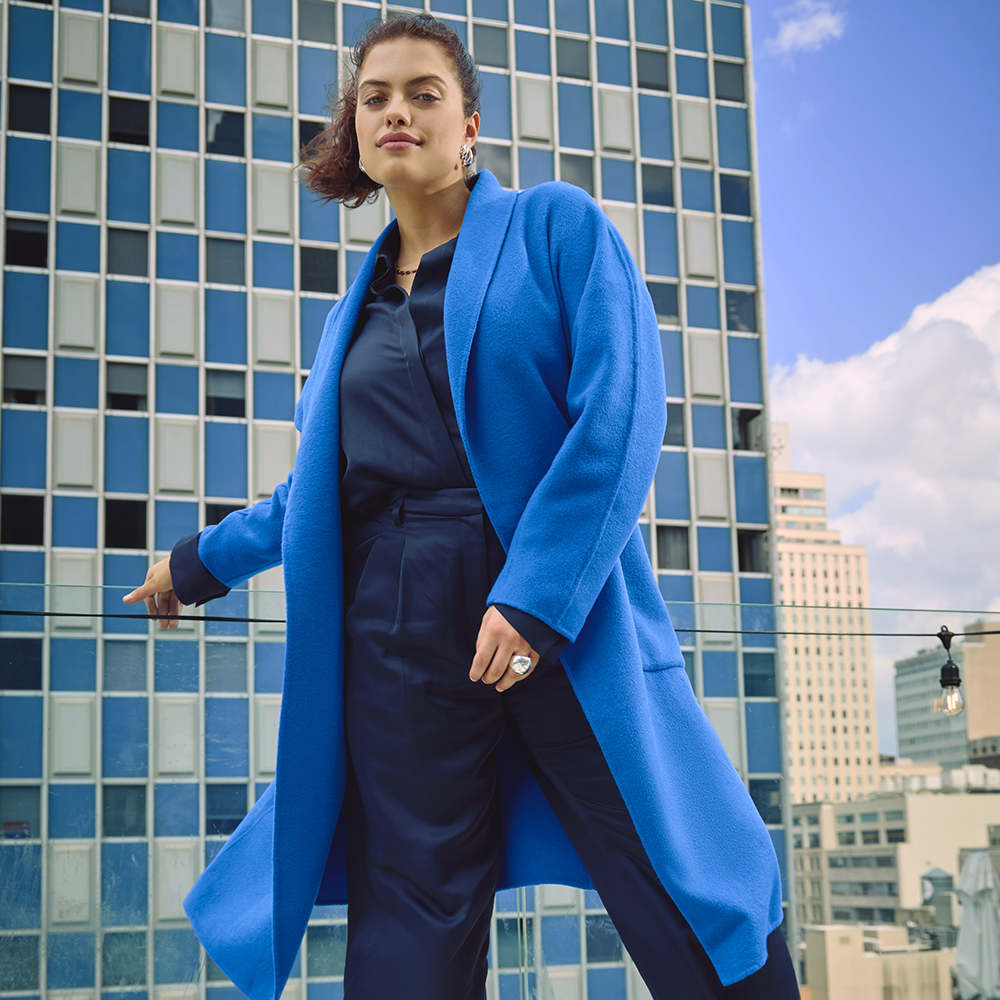 Model with the cobalt coat standing in front of a cityscape with a blue sky.